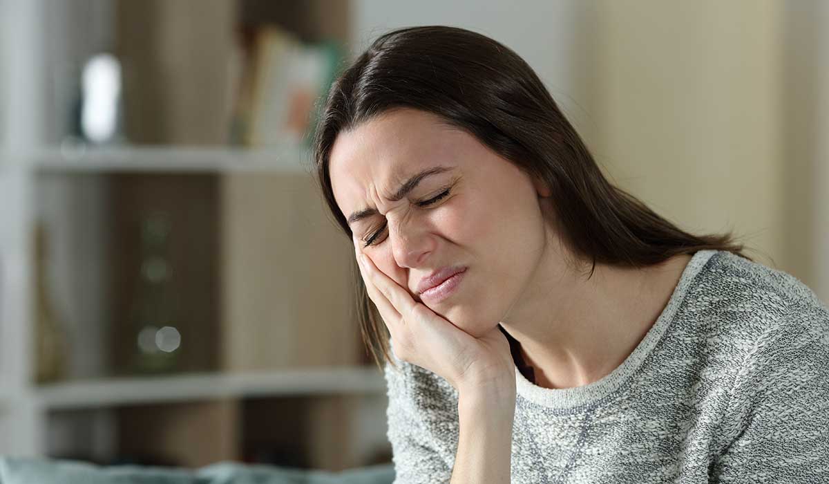 A woman sits indoors with her eyes closed, holding her cheek and wincing in pain, suggesting she has a toothache or facial discomfort.