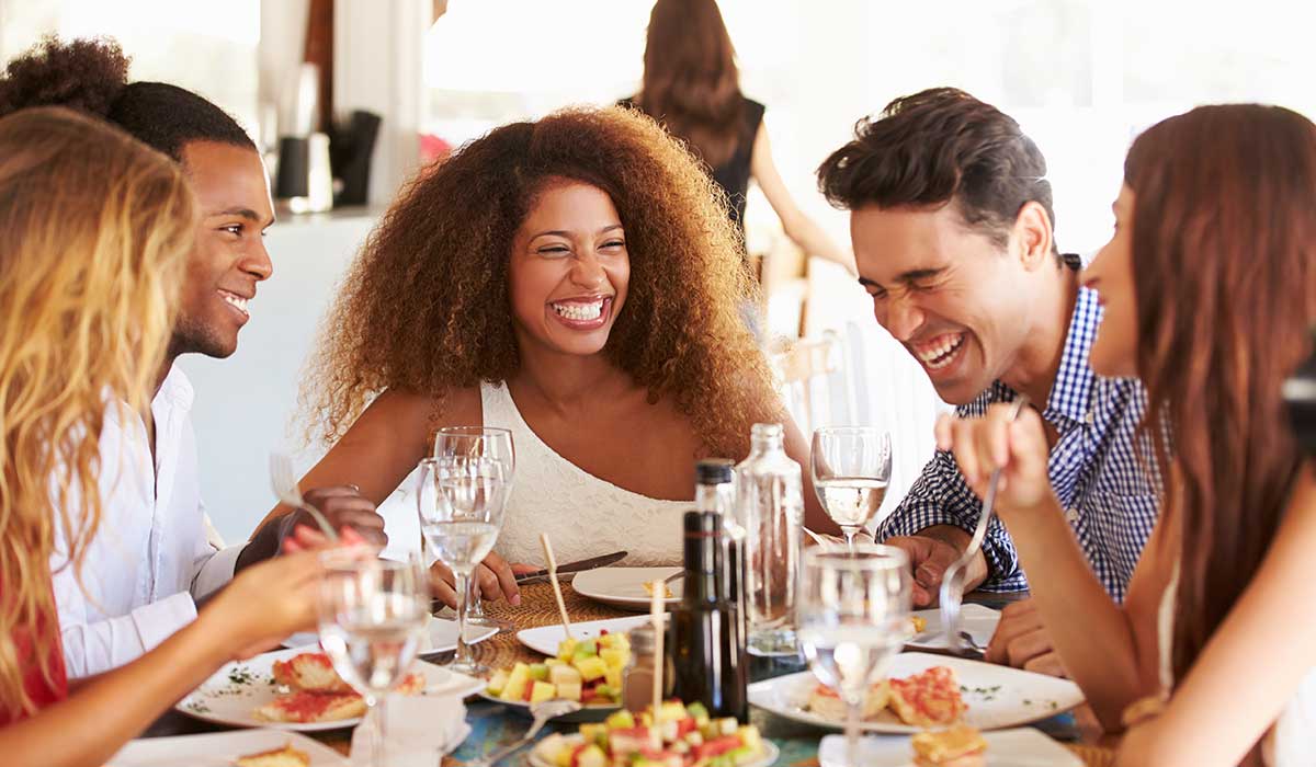 A group of young adults sit around a table, laughing and enjoying a meal together. The table is set with plates of food, glasses of water and wine, and everyone appears to be having a good time.