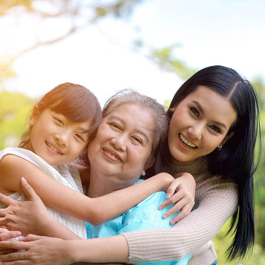 Three generations of women—child, elderly woman, and adult—embrace each other and smile warmly outdoors, with trees and soft sunlight in the background.