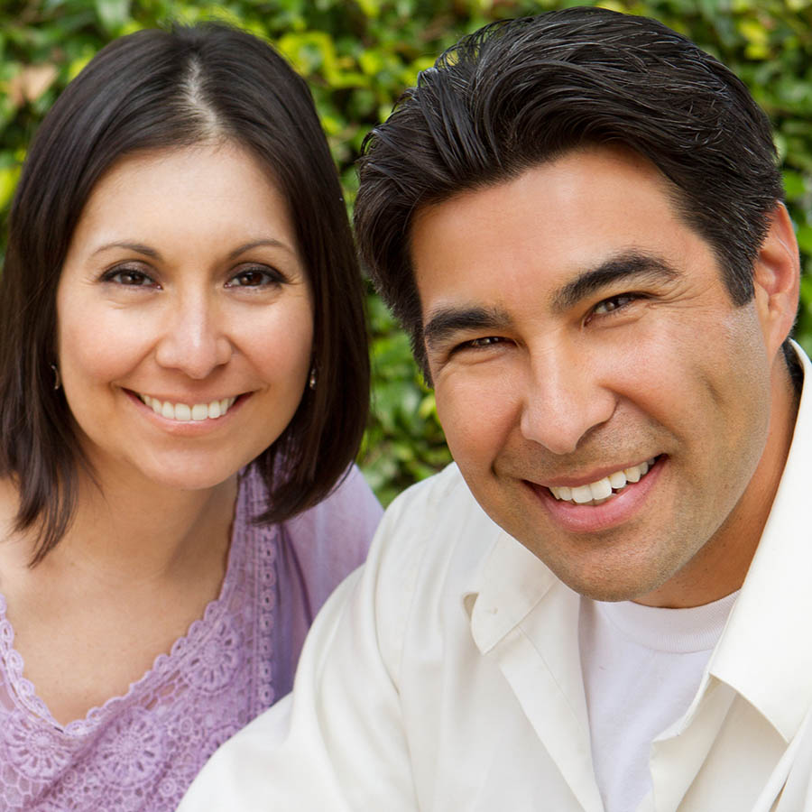 A smiling man and woman pose closely together outdoors, with greenery in the background. The woman has shoulder-length dark hair and wears a lavender top; the man has short dark hair and wears a white shirt.