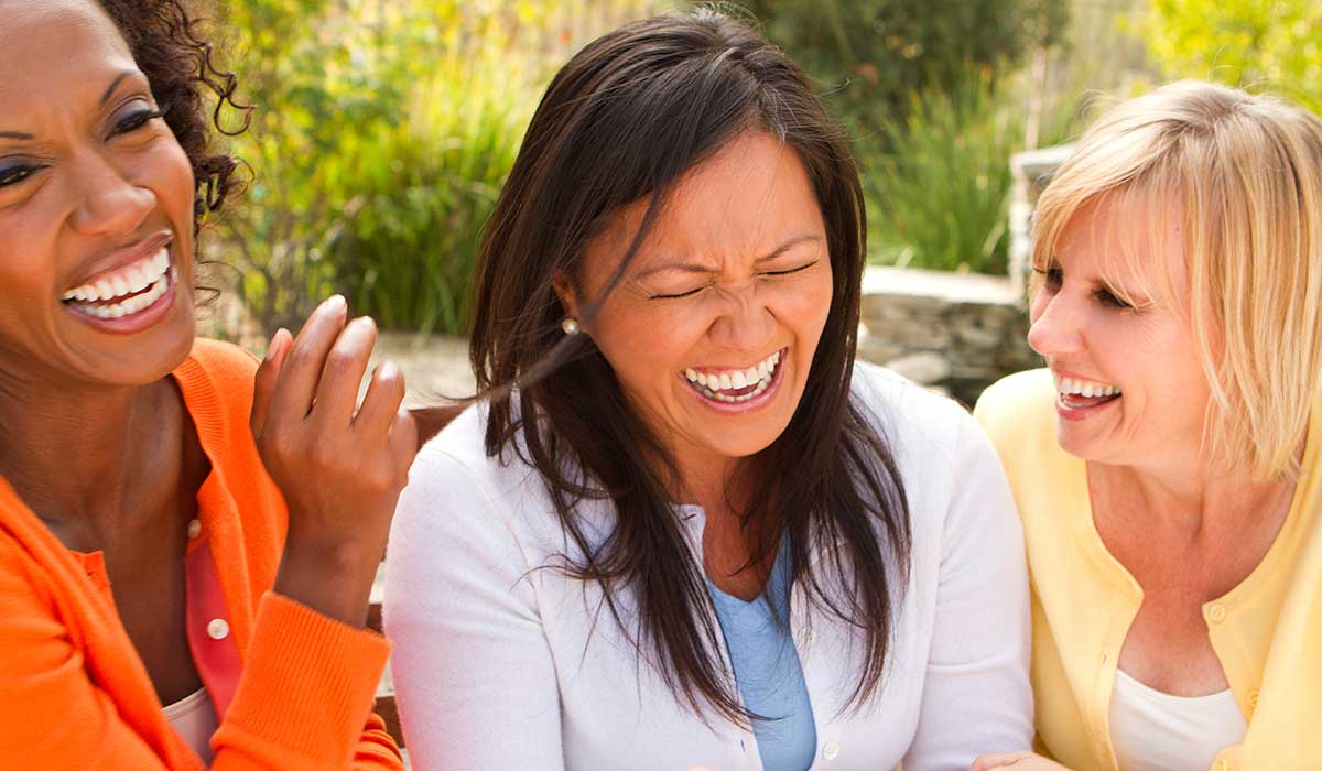 Three women sit close together outside, laughing joyfully. They appear to be friends enjoying each others company, dressed in colorful sweaters, with greenery visible in the background.
