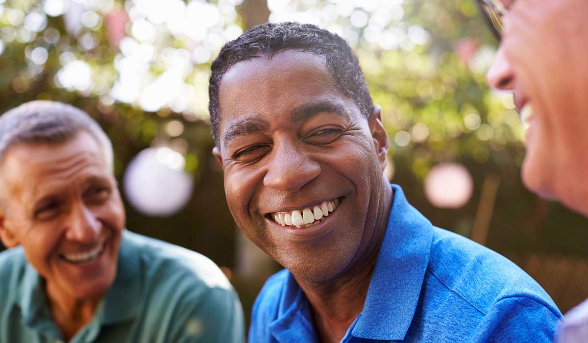 Three men sit close together outdoors, smiling and laughing. The man in the center, wearing a blue shirt, is the focus, while the other two men are partially visible, engaged and joyful. Sunlight filters through trees in the background.