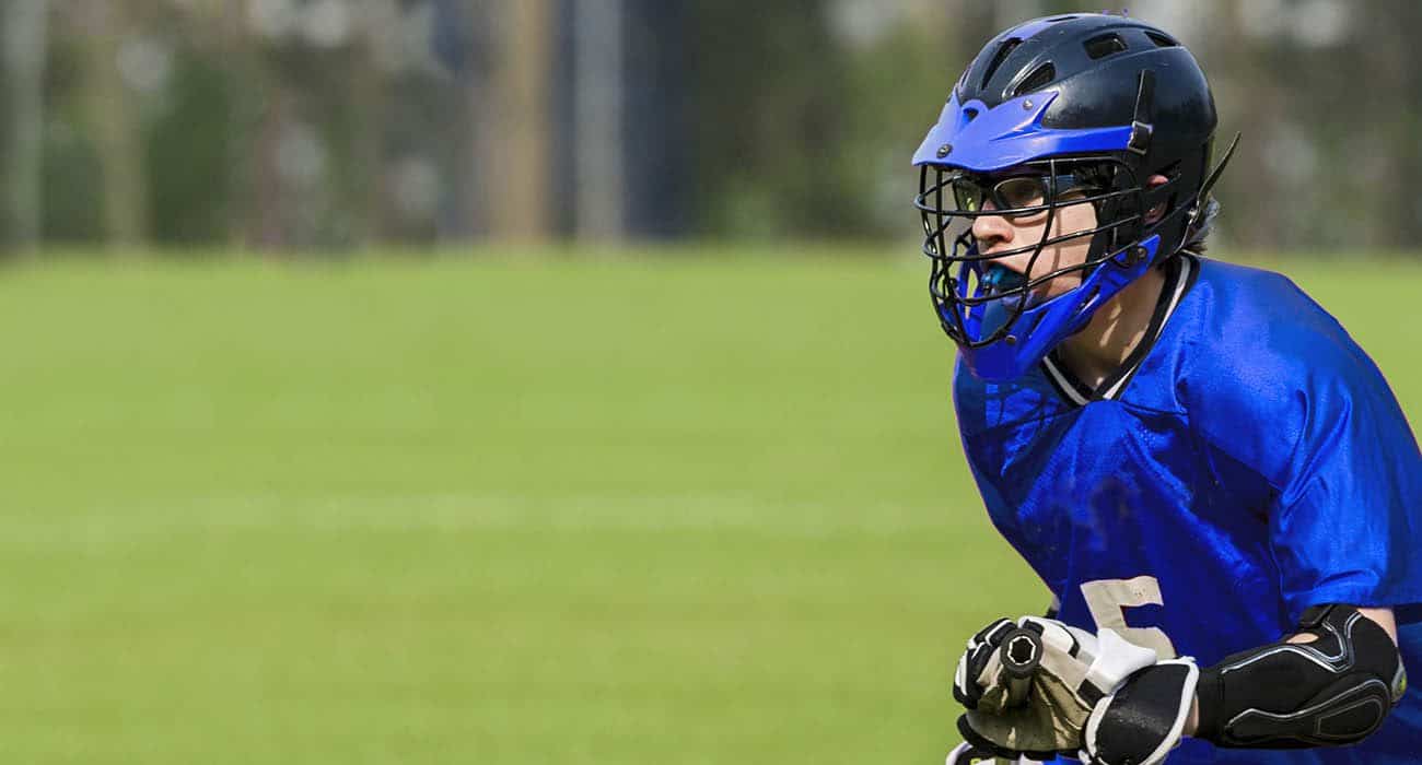 A lacrosse player in a blue jersey and black helmet crouches in an athletic stance on a grassy field, ready for action.